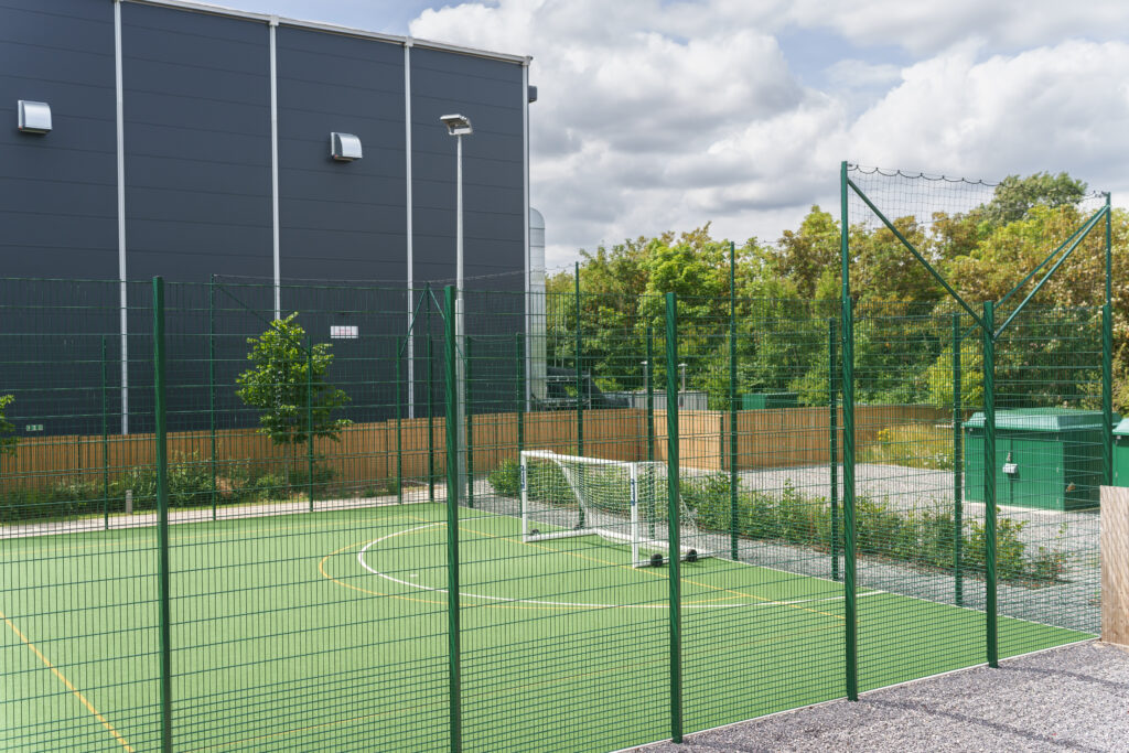 Outdoor fenced sports court with green surface, a small football goal, and netting; modern building and trees in the background under a partly cloudy sky.