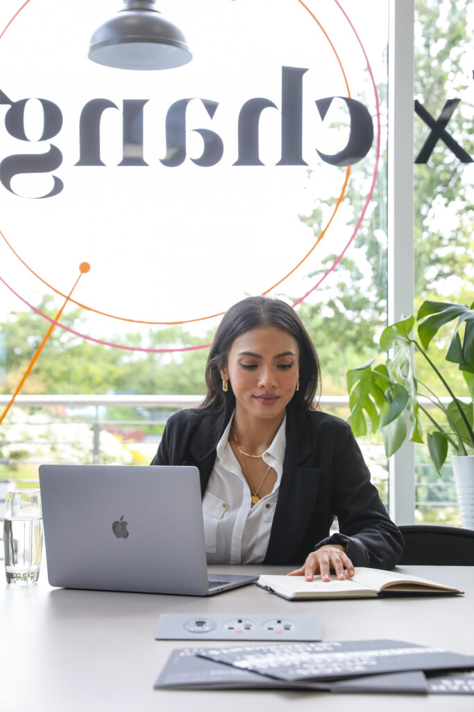 A woman in business attire sits at a desk with a laptop and notebook, working in a modern office with large windows and greenery outside.