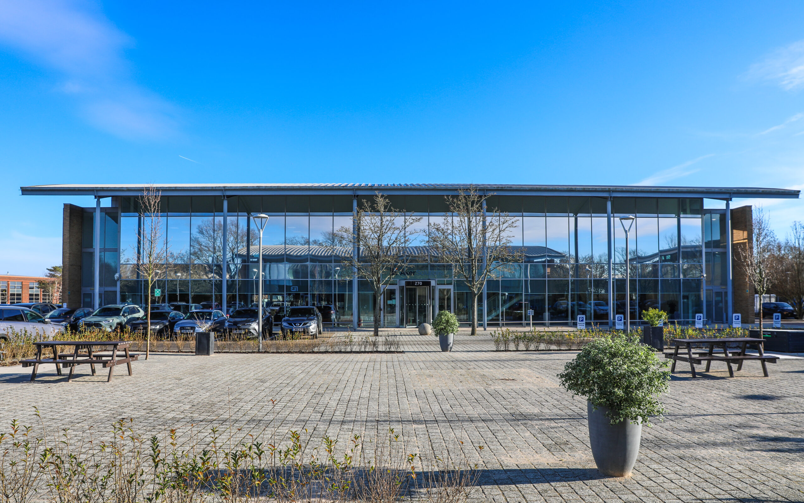 Modern glass-fronted building with large windows, several cars parked in front, benches, and potted plants on a paved plaza under a clear blue sky.