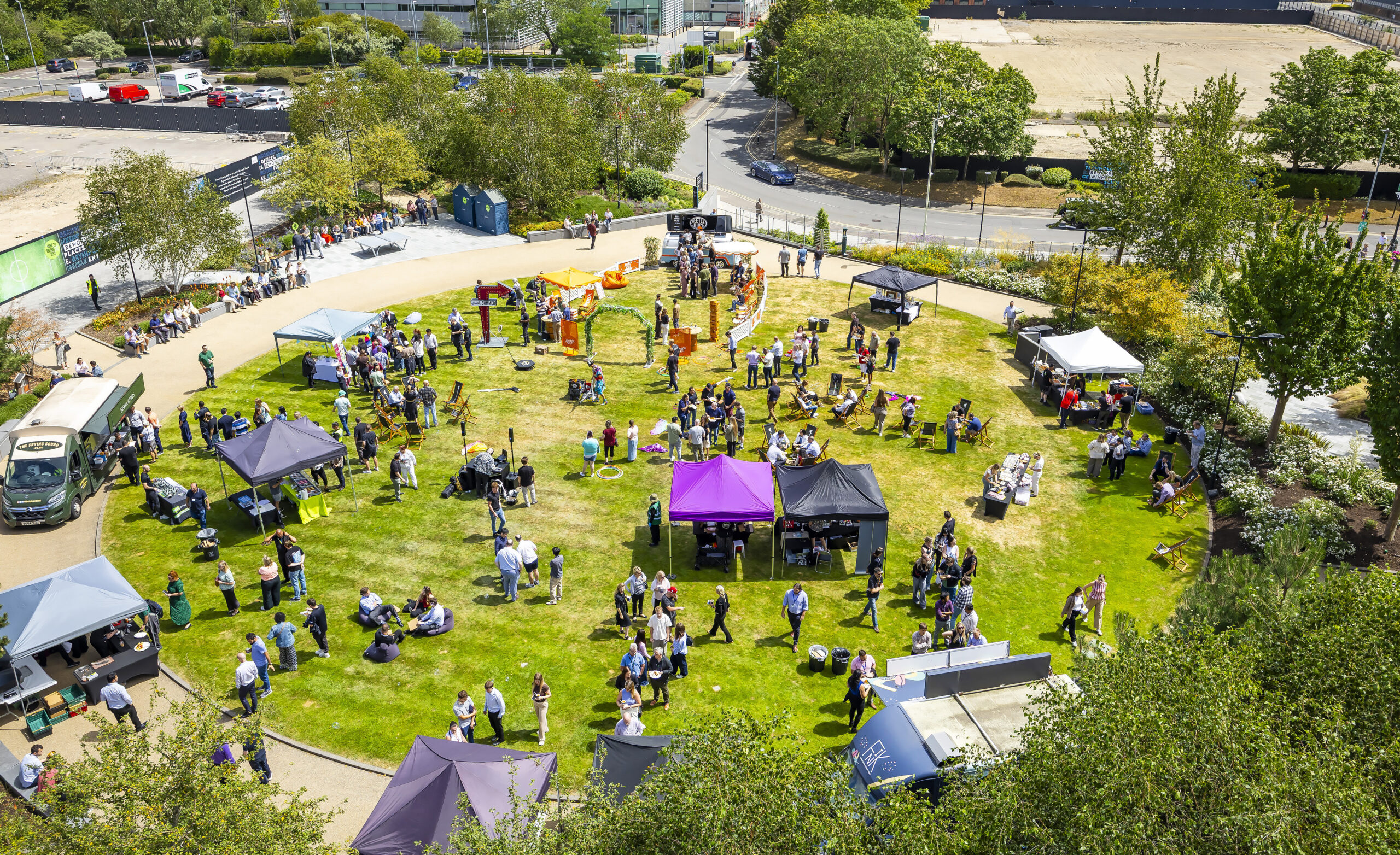 Aerial view of an outdoor event with people, marquees, and tables arranged in a circle on a grassy area surrounded by trees and roads.