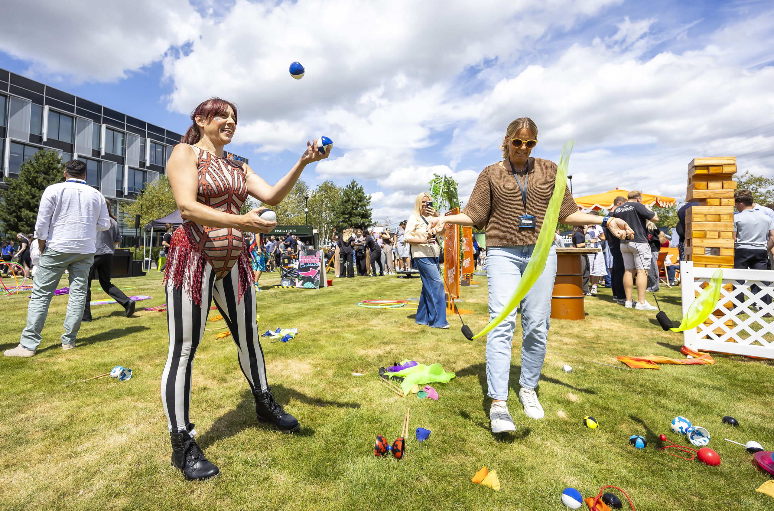 Two women juggle and perform tricks with props on a grassy lawn during an outdoor event, with scattered toys, games, and a crowd in the background.