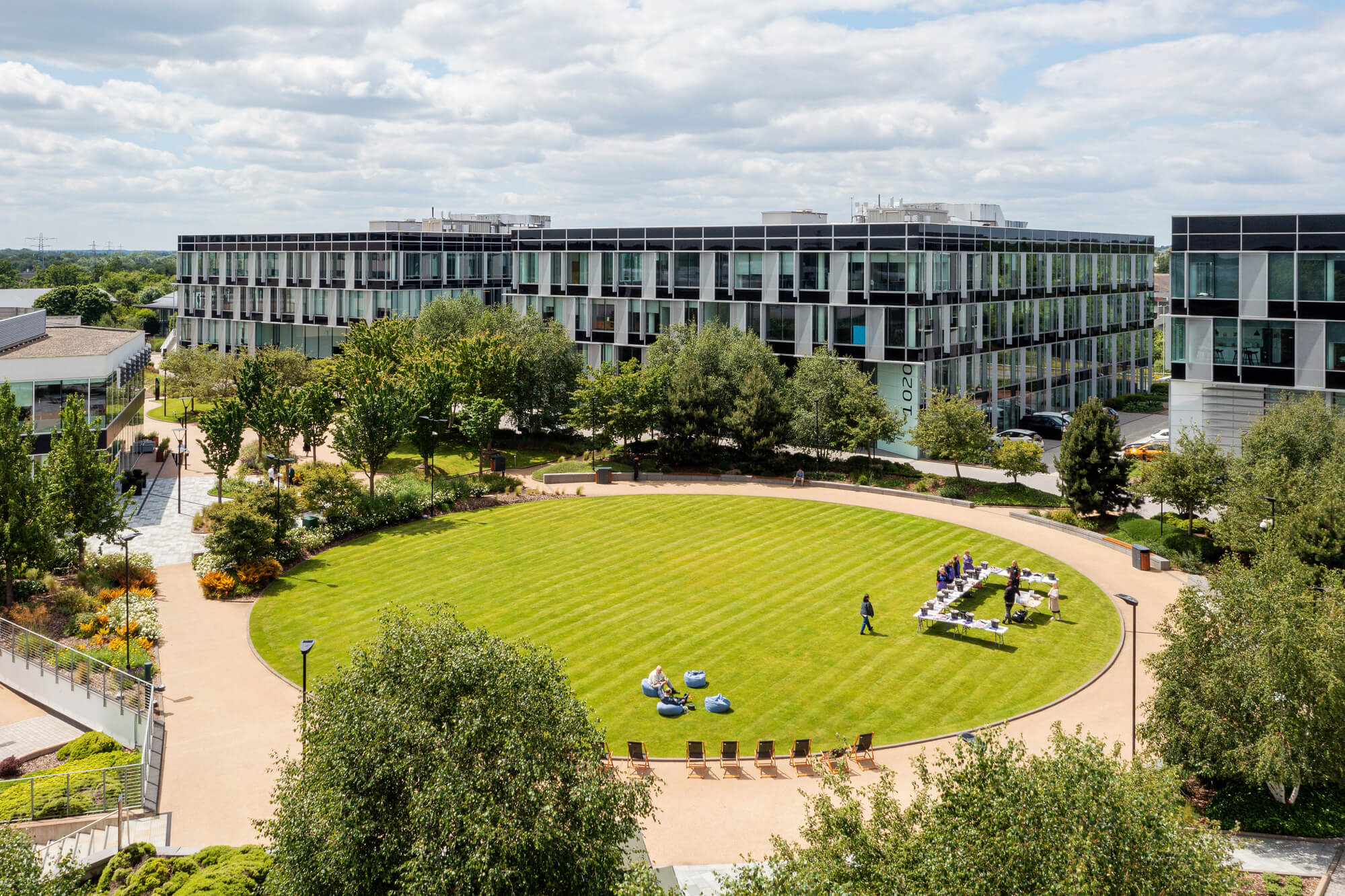 A modern office complex with glass buildings surrounds a circular green lawn, where people are gathered and chairs are arranged on the grass.
