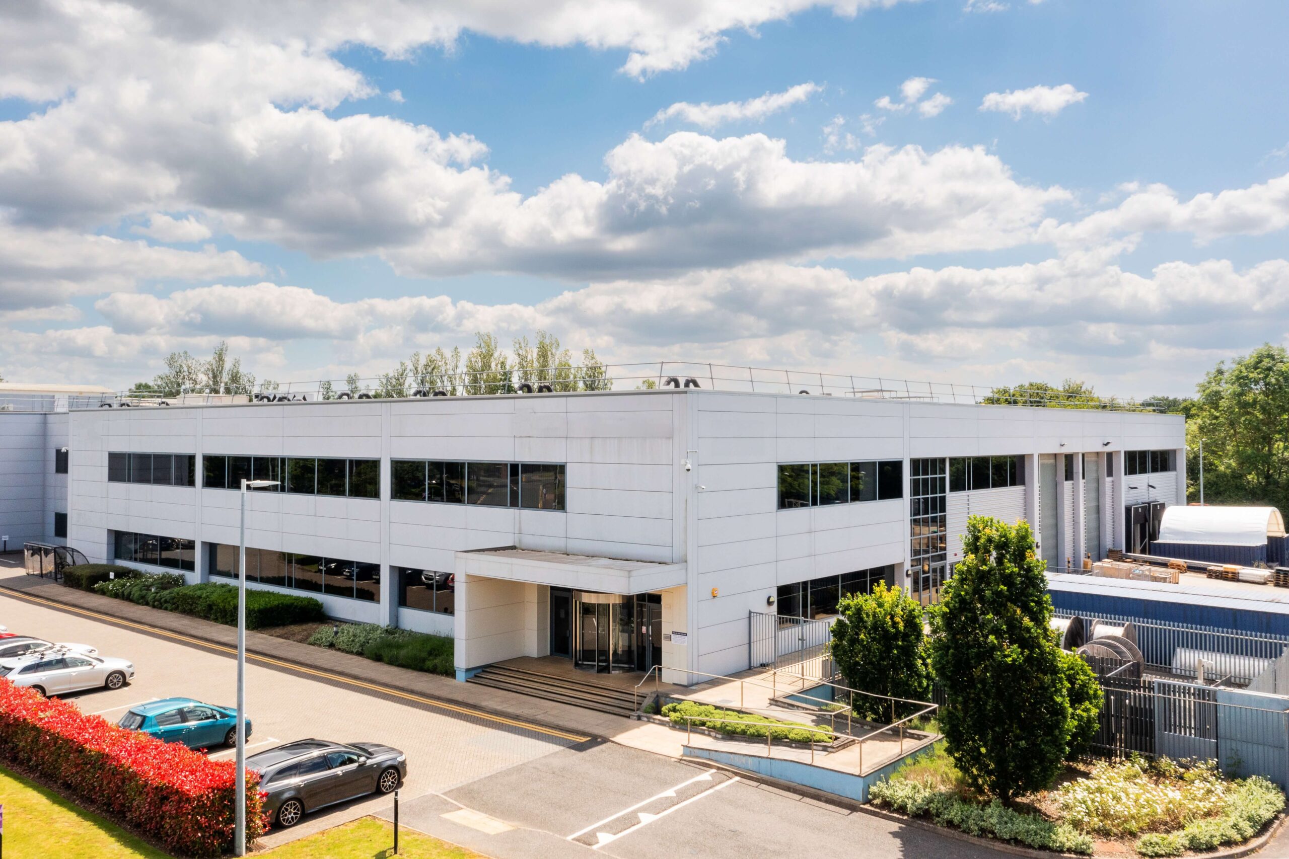 A modern white industrial building with large windows, surrounded by a car park with parked cars, trees, and a partly cloudy sky above.