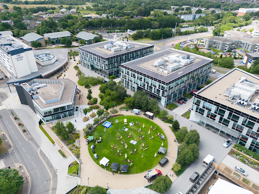 Aerial view of modern office buildings surrounding a circular green lawn with people sitting and several food vans nearby. Trees and roads are visible in the background.